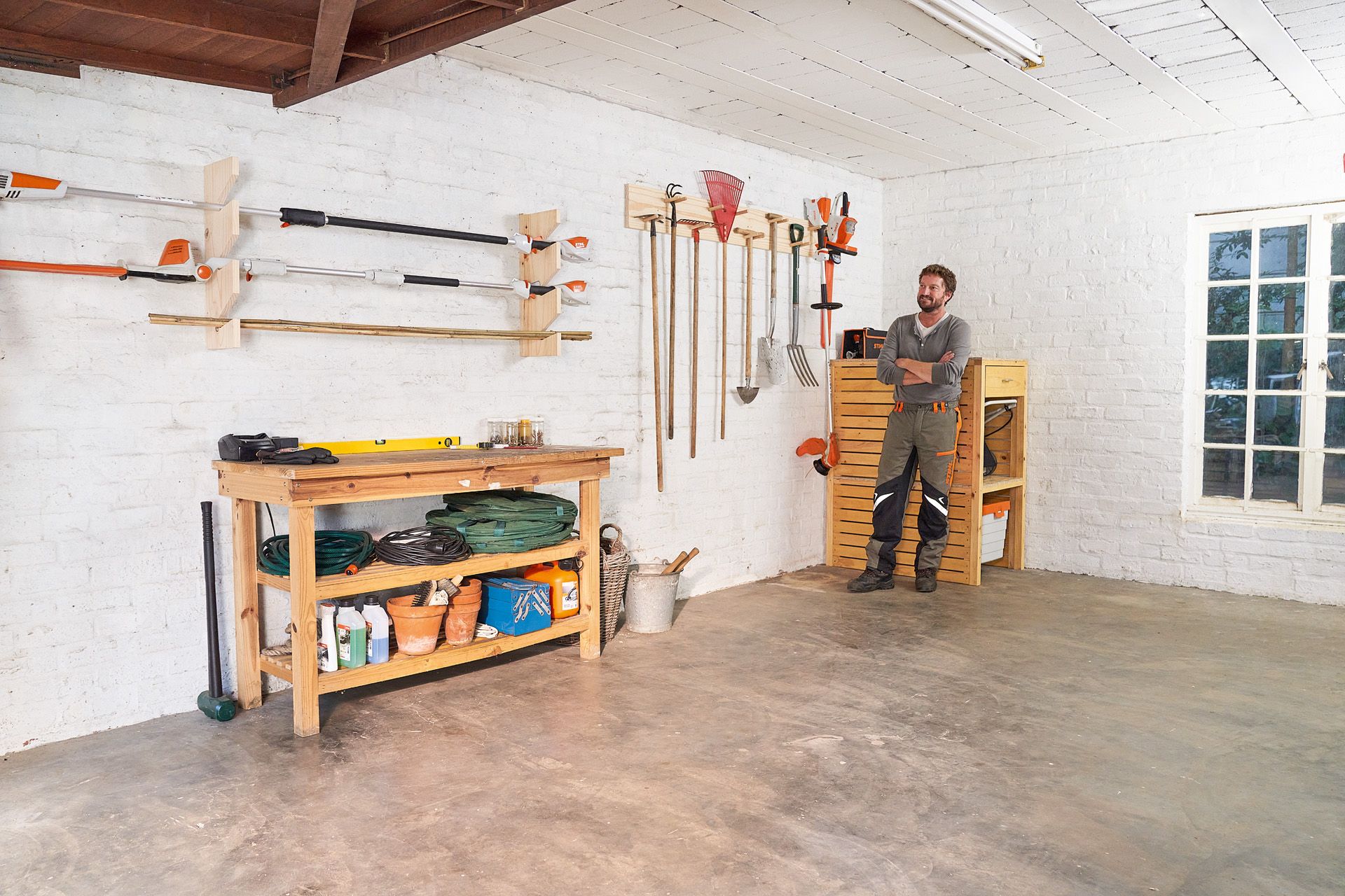 Um homem de braços cruzados apoiado num suporte para ferramentas e máquinas de jardinagem numa garagem 